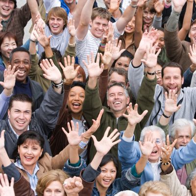 A photograph taken from above of a crowd of people of mixed ages, races and genders, all looking up at the camera and smiling and holding their hands in the air